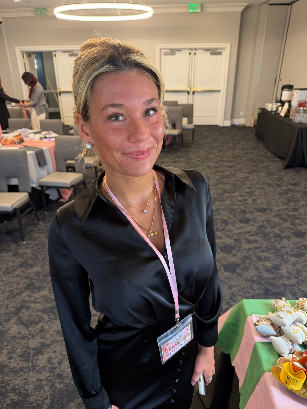 A woman with blonde hair tied back, wearing a black satin long-sleeve shirt and a pink lanyard with an ID badge, standing in a conference room setting.