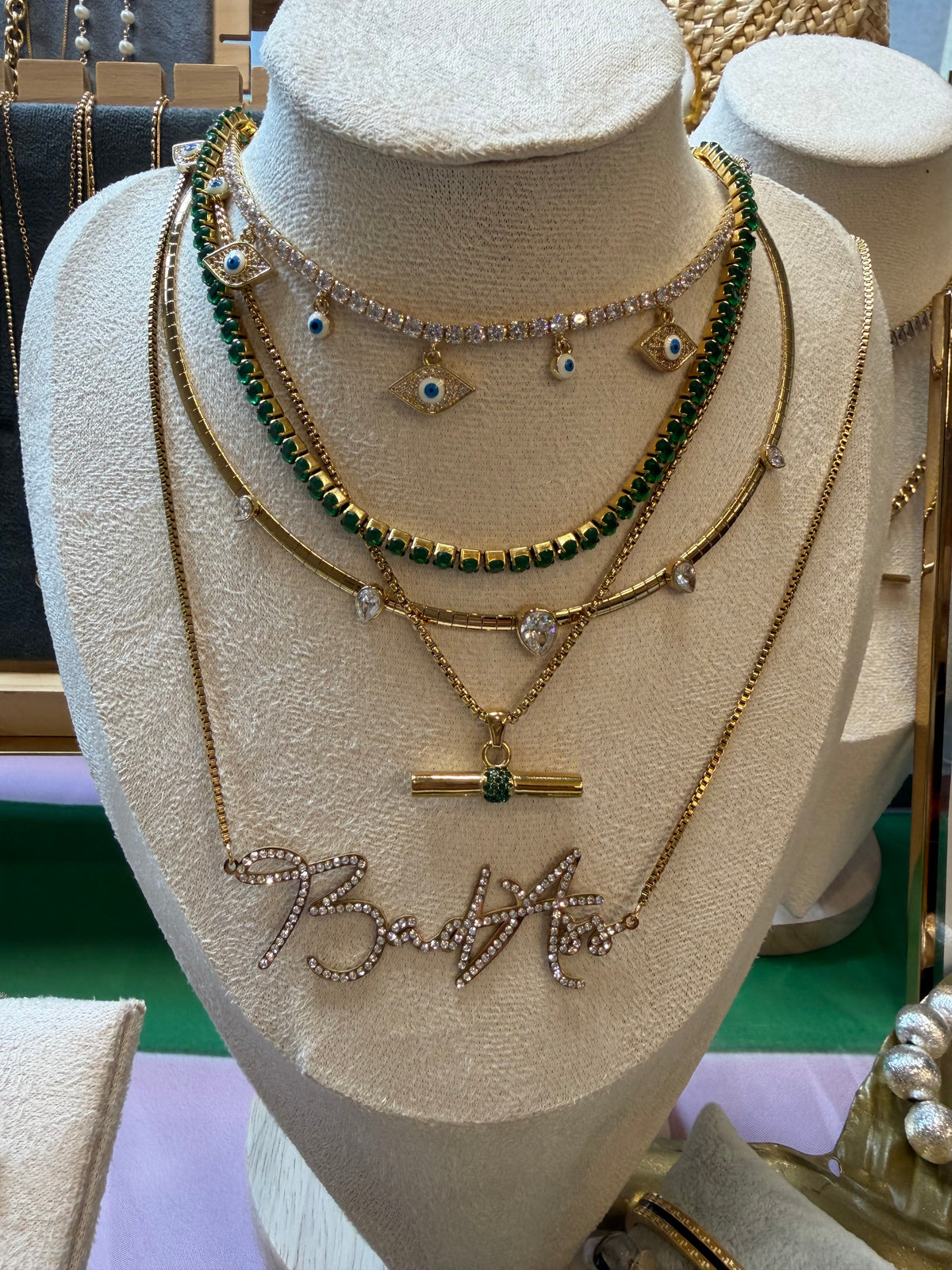 A layered necklace set displayed on a cream fabric mannequin bust. The set includes a gold chain with a rhinestone script pendant spelling 'BadAss', a gold chain with a small gold bar and green stone accent, a gold chain with evil eye charms, a green beaded chain, and a gold chain with clear rhinestone eyelets.