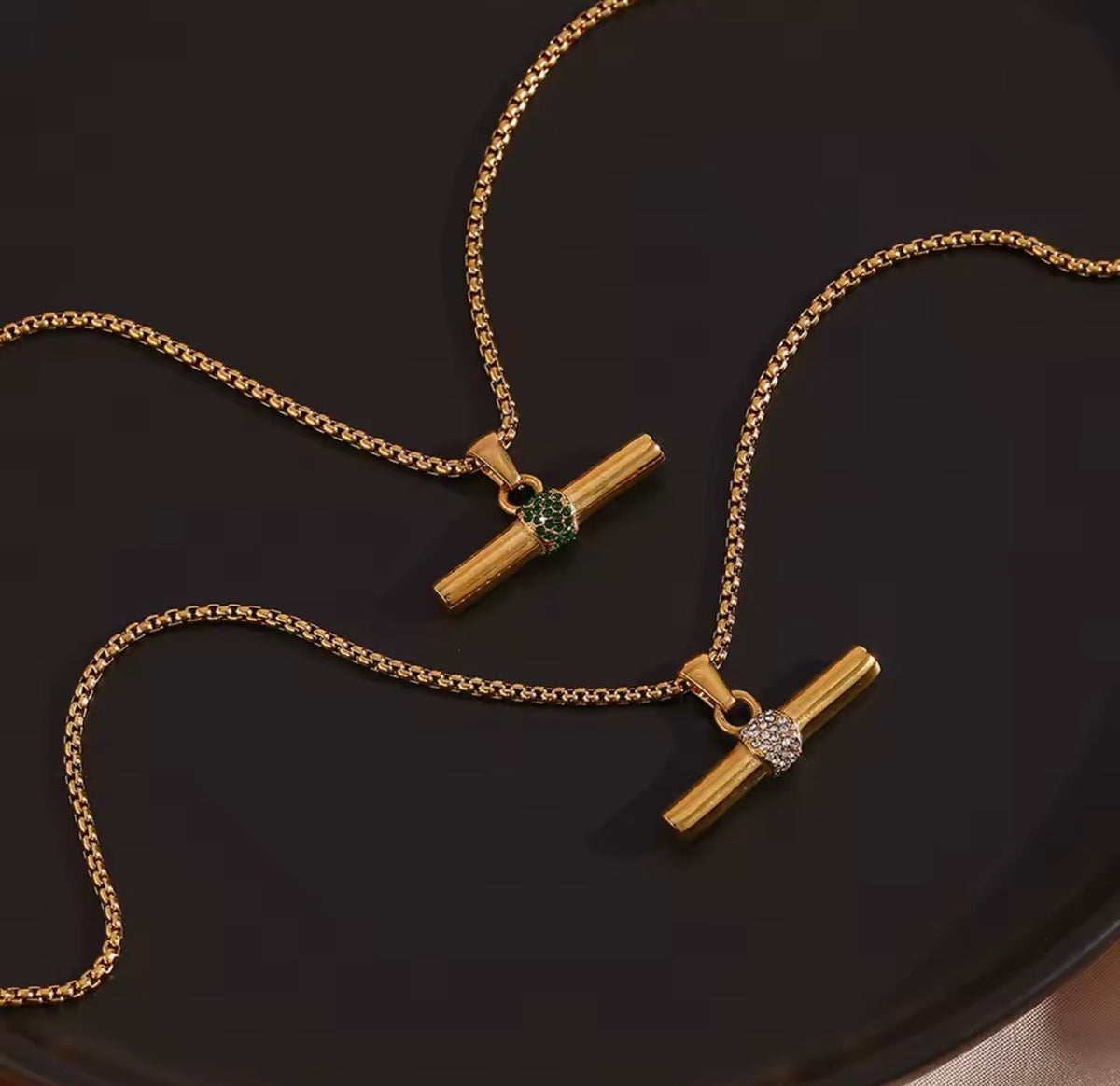 Two gold-toned necklace pendants with cylindrical bar charms and small round stone accents, one green and one clear, displayed on a dark surface.